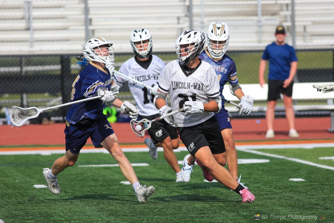 Lincoln-Way lacrosse player #21 with pink cleats cradling ball vs navy defender
