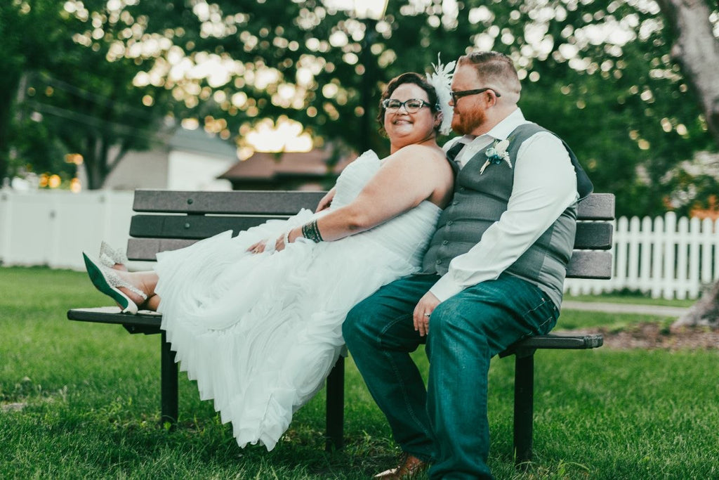 Bride and groom sitting on park bench — she in white tiered dress, he in grey vest and jeans