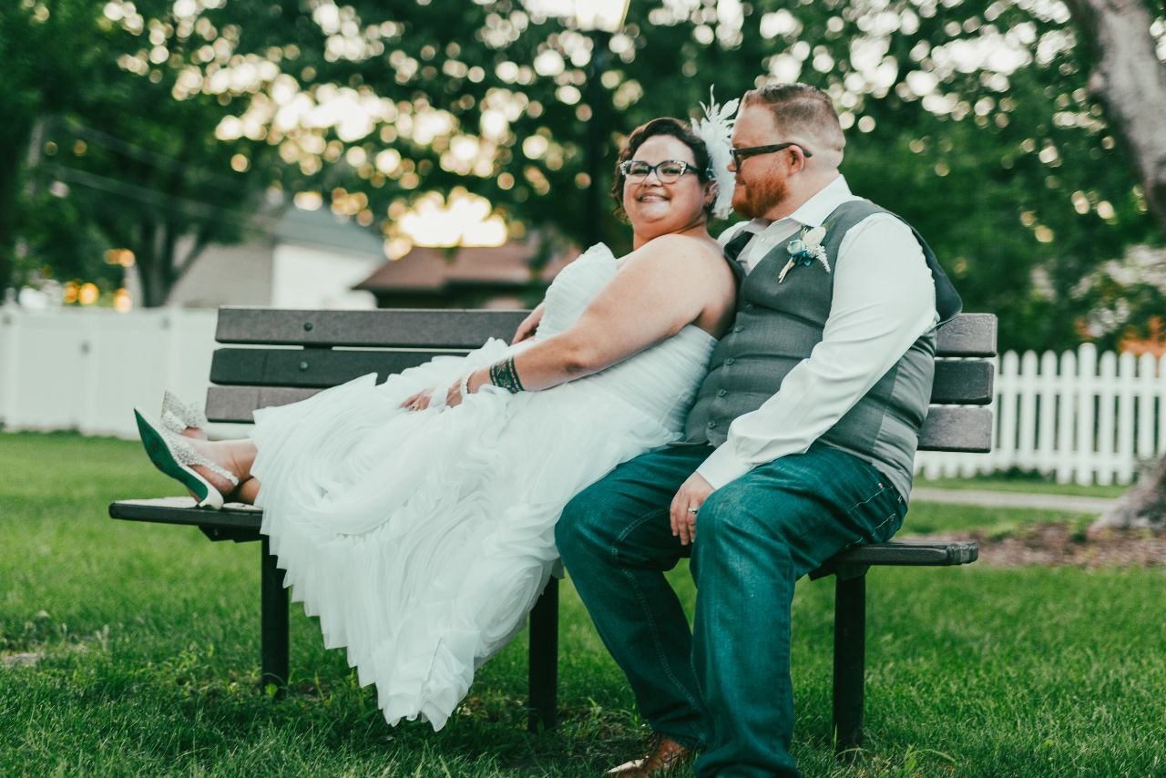 Bride and groom sitting on park bench — she in white tiered dress, he in grey vest and jeans
