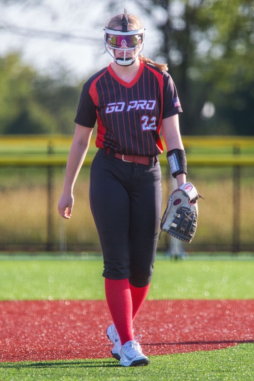 Softball player in black GO PRO jersey walking baseline, helmet with pink goggles